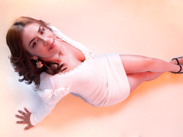 Theresa Larson, acostada de lado, vestida con un vestido blanco de manga larga, que muestra sus pechos y glúteos, con cabello castaño rizado, tacones negros y aretes blancos, sobre un fondo color melocotón..
