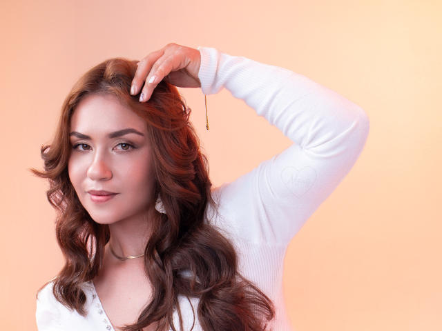 Theresa Larson, com cabelo castanho ondulado, camisa branca de manga comprida, mão no cabelo, brinco, sorriso discreto, batom rosa, fundo rosa suave, brincos brancos, maquiagem suave, pose relaxada..