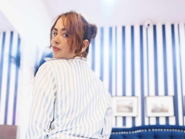 TheresaLarson with brown hair in a messy bun, wearing a blue and white striped shirt, looking over her shoulder with a slight smile, in a brightly lit room.