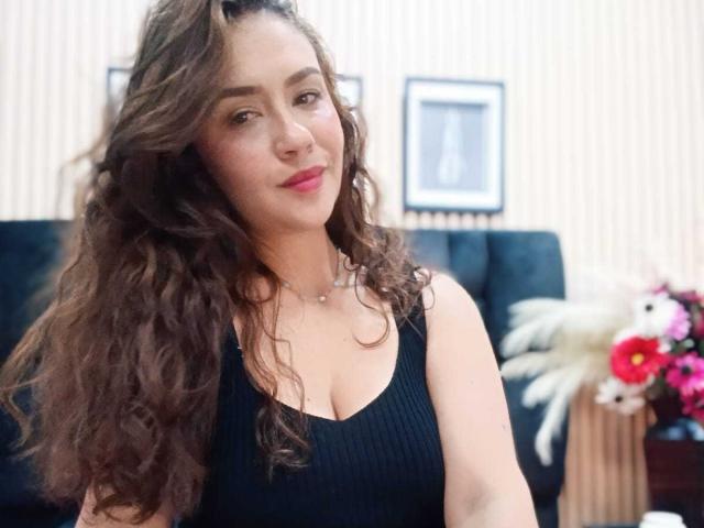 Theresa Larson, con cabello castaño rizado y largo, vestida con una camiseta negra de tirantes con textura, mostrando sus senos, con lápiz labial rosa y una sonrisa sutil, en una habitación bien iluminada con flores..