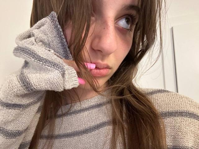 KarieMestler with brown hair, pink lipstick, and a grey striped sweater, looking to the side with a slightly pouty expression, hand touching her face.