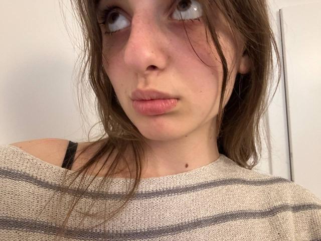 KarieMestler's pouty face with slightly parted pink lips, brown eyes looking upwards, messy brown hair, beige striped sweater, black bra strap visible, fair skin with small moles.