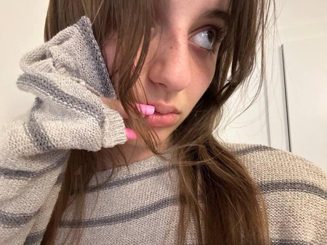 KarieMestler with brown hair, pink lips, and a grey striped sweater, biting her pink-manicured finger, looking to the side with a slightly pouty expression.