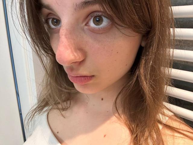 KarieMestler's face with brown eyes, slightly freckled skin, and small nose, wearing a white top, brown hair, and a neutral expression. Her tits are not visible.