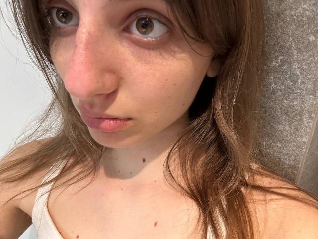KarieMestler's face showing brown eyes, pink nose, and slightly parted pink lips. She has long brown hair, freckles, and is wearing a white tank top. Her ass is not visible.