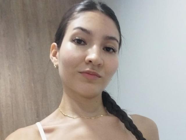LucianaBetancour has dark brown hair in a braid, wearing a white tank top, gold necklace, and earrings, with a slight smile and clear skin. Her tits are small.