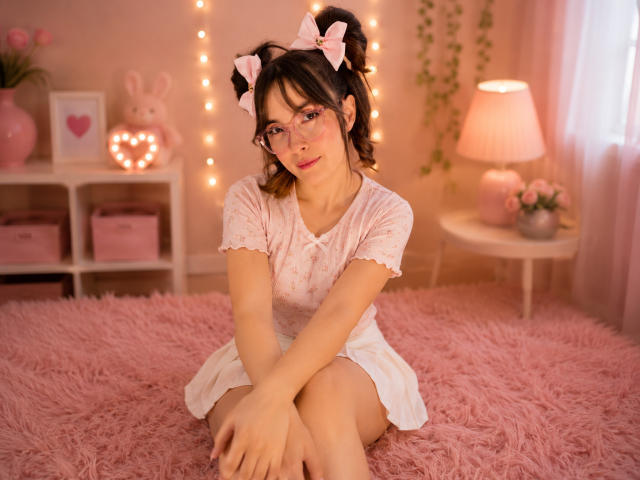 IsabelaP with cute pink hair, glasses, and pink bows, sitting on a pink fluffy bed, wearing a white polka-dot dress, looking shy with her legs crossed.