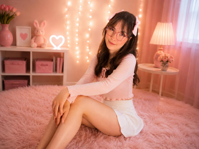 IsabelaP with long dark hair, pink glasses, pink bow, pink top, white skirt, sitting on pink fluffy bed, legs together, cute, pink fairy lights, pink room, smiling.