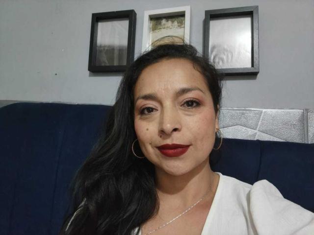 StangelarRivera has dark hair, red lipstick, hoop earrings, a white shirt, and a slight smile, with a small mole on her right cheek, looking directly at the camera.