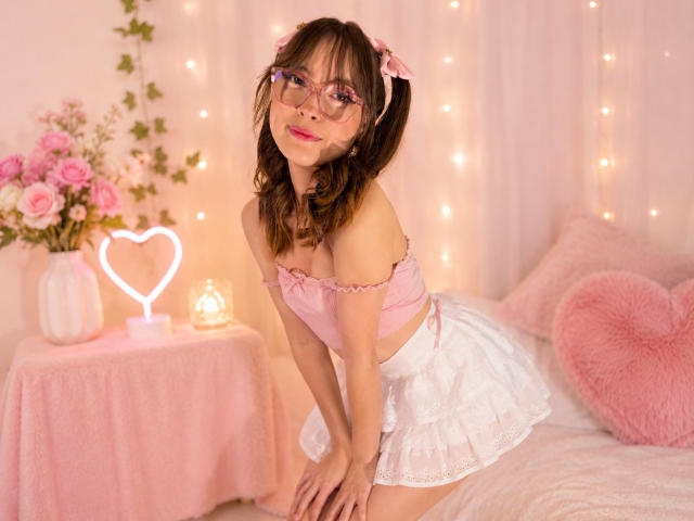 IsabelaP with pigtails, pink glasses, pink off-shoulder top, white lace skirt, pink bow, leaning forward on bed, pink heart lamp, pink flowers, and fairy lights in the background.