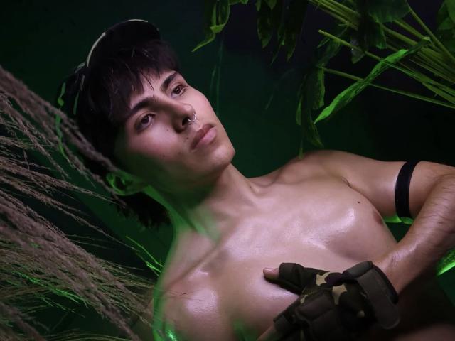 PepperTwink, torse nu, avec une peau brillante et moite, des gants noirs, un anneau au nez, et une lumière verte, regardant vers le haut, avec des plantes et de l'herbe au premier plan..