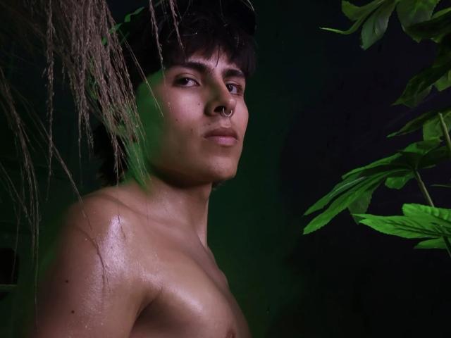 PepperTwink a une peau brillante et humide, de couleur tan, des cheveux foncés et en désordre, un anneau de nez et un regard sérieux. Des plantes vertes et feuillues se trouvent sur la droite. PepperTwink est torse nu..