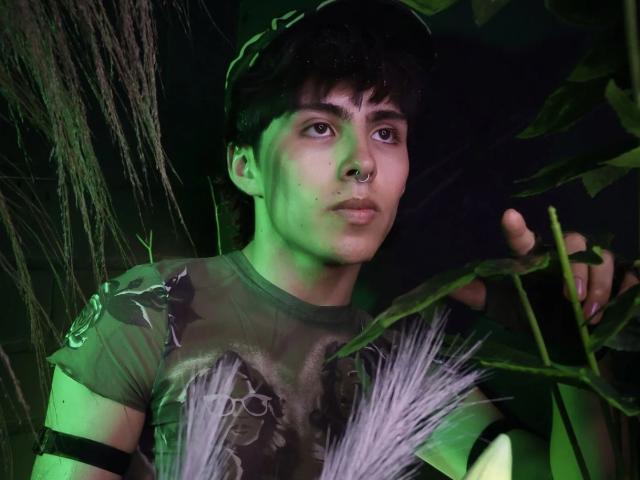 PepperTwink, avec ses courts cheveux noirs, un anneau au nez, un visage légèrement verdâtre, une chemise de camouflage, tenant une plante, lumière verte, expression sérieuse, bracelet à l'avant-bras, feuille au premier plan, légèrement flou..