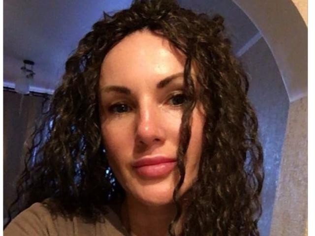 XMelanieWomans hat lockiges, braunes Haar, helle Haut und rosafarbene Lippen mit einem leichten Lächeln. Sie trägt ein braunes Hemd, und ihre blauen Augen schauen in die Kamera..