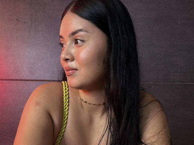 AlannaVelvet with long black hair, glossy pink lips, light brown skin, green strap, subtle makeup, and a thin necklace, looking to the side with a slight pout.