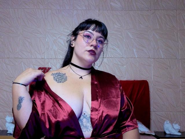 NaniSky with glasses, black choker, and red satin robe, showing her tits, tattoos on chest and arm, and a bracelet, sitting against a textured wall.