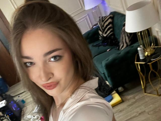 ScarletShow has light brown hair, green eyes, and red lipstick. She's wearing a white top, looking at the camera with a slight smile, in a cozy room.