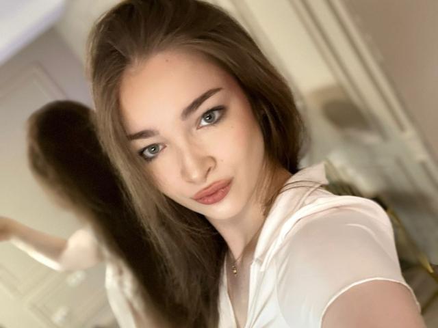 ScarletShow has green eyes, brown hair, and is wearing a white shirt. Her reflection is visible in the mirror behind her. She's looking at the camera with a subtle pout.