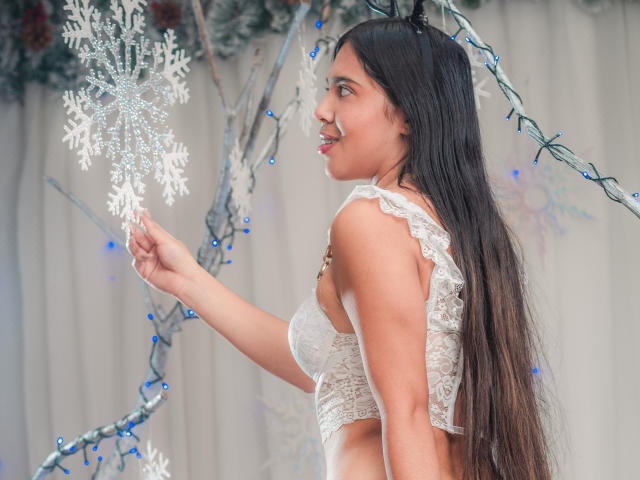 VioletaBlis with long black hair, white lace bra, holding a silver snowflake, smiling, standing side profile, looking right, wearing a necklace, blue Christmas lights in background.