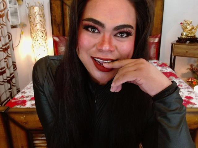 TatianaCocker with dark hair, red lipstick, and long eyelashes, biting her finger, wearing a black leather jacket, smiling at the camera, showing off her sexy, full lips.