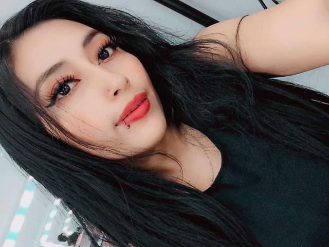 AilenRosse with long black hair, red lips, and a lip piercing, wearing black, showing off her cute face and sexy makeup, looking at the camera with a slight smile.