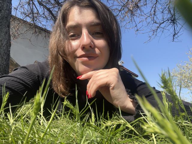 Baby2223 con capelli castano di media lunghezza, maglietta nera a coste, unghie rosse, sdraiata sull'erba, giornata di sole, cielo blu, sorridente, mano sotto il mento, rami di alberi sullo sfondo..