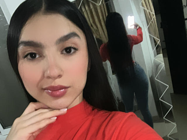 MilanMoon with long black hair, red turtleneck, holding phone, reflecting in mirror, showing her ass in tight jeans, smiling, hand on cheek, glossy pink lips, in selfie.