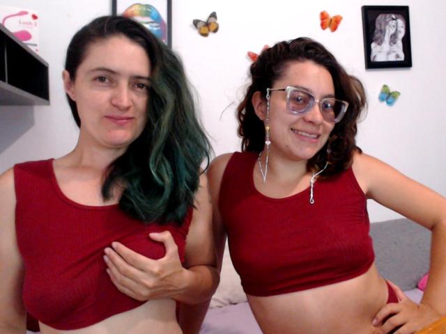 LunaVenus met groen uitlopend haar en LunaVenus met krullend bruin haar en een bril, beiden met rode crop tops die hun borsten bedekken, lachend in een slaapkamer..