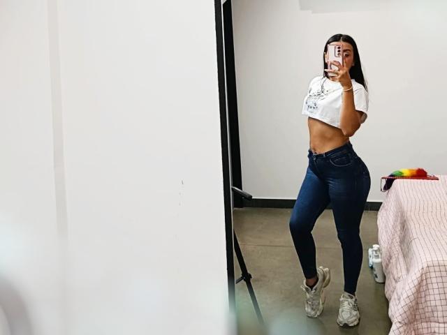 ZammySanz en un selfie frente al espejo, vistiendo una camiseta corta blanca, jeans azules de cintura alta y zapatillas blancas, mostrando un trasero curvilíneo y tonificado y pechos pequeños y firmes..