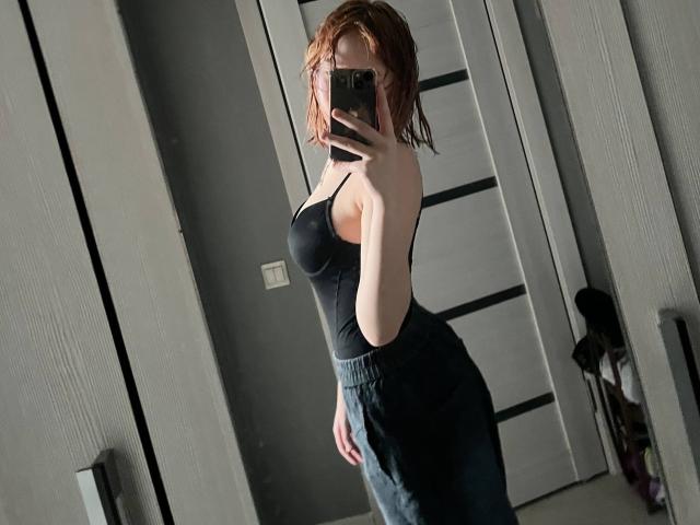 Margeret Eskuchen, met nat rood haar, een zwart tanktopje dat haar borsten laat zien, en donkergrijze broek, maakt een spiegelzelfportret met haar telefoon, waarbij haar billen in beeld zijn..