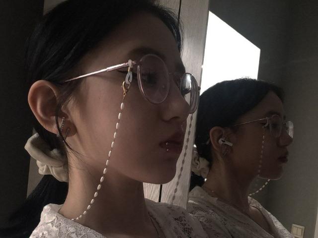 Margeret Eskuchen, com óculos redondos, colar de pérolas e piercing no lábio, refletida em um espelho, com cabelo preto preso em um elástico branco, vestindo uma blusa de renda, em um quarto escuro, usando fones de ouvido..