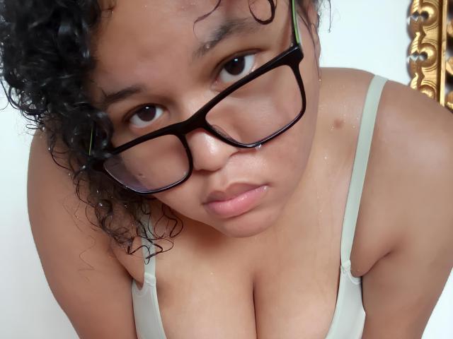 KuroNyG met krullend zwart haar, een bril en een groen tanktop, waarbij haar natte borsten zichtbaar zijn en ze een serieuze uitdrukking heeft, terwijl ze naar beneden kijkt in de richting van de camera..