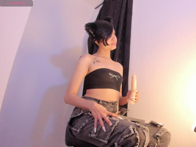CamilaLadySmith mit schwarzem Oberteil, kleinen Brüsten, Tattoo auf der Schulter, hält einen großen Dildo, trägt zerrissene Jeans, sitzt auf einem Hocker, schwarzes Haar im Dutt, lächelt..