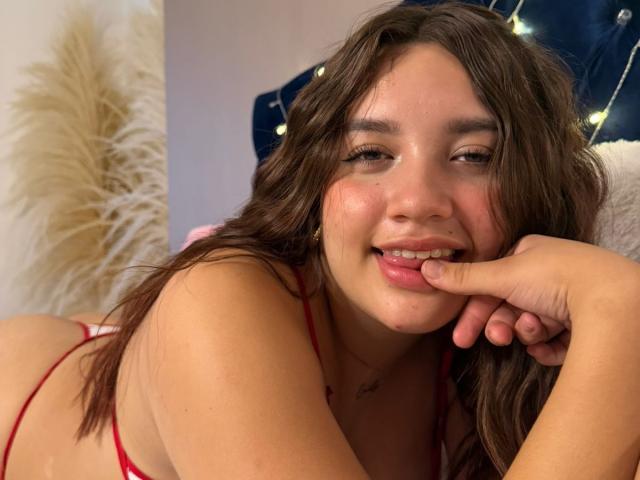 SofiMillerr with brown wavy hair, red lingerie, smiling with finger in mouth, showing her cute, chubby face, and her red thong-covered ass, lying on a bed.