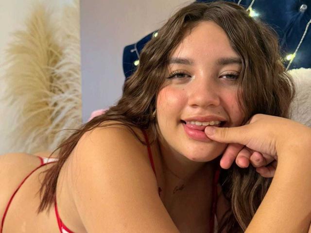SofiMillerr, with long brown hair, red lingerie, and a cute smile, bites her finger while lying on a bed, her ass and tits visible, looking sexy and relaxed.