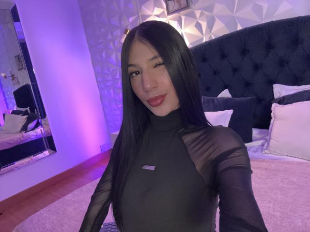 Sophie Beaumont, avec de longs cheveux noirs, clignant de l'œil, portant un haut noir transparent qui révèle ses petites seins, avec du rouge à lèvres, dans une chambre avec un éclairage violet et une tête de lit bleu foncé capitonnée..