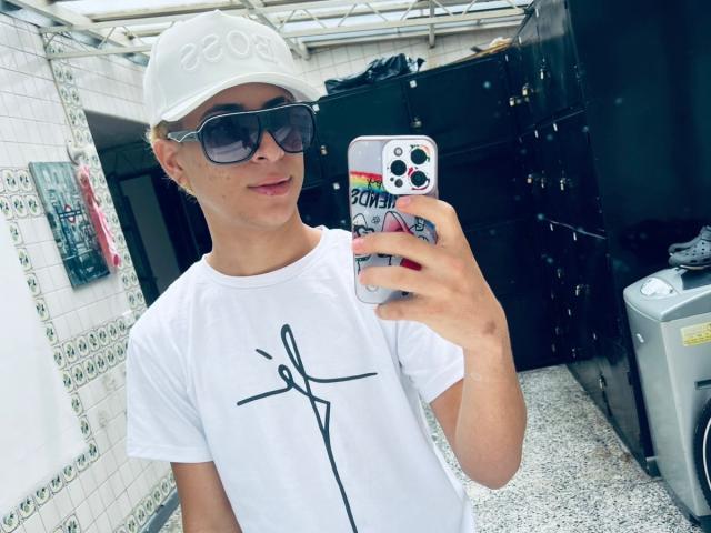 HollmanFronsh, usando um boné branco, óculos de sol e uma camiseta branca com um desenho em preto, está tirando uma selfie em um banheiro com armários pretos. O celular dele tem uma capa colorida..