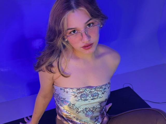 ElodieGardner with glasses, blonde hair, and a strapless, floral-patterned dress, sitting, showing her small tits and a cute face with a slight smile, under blue lighting.