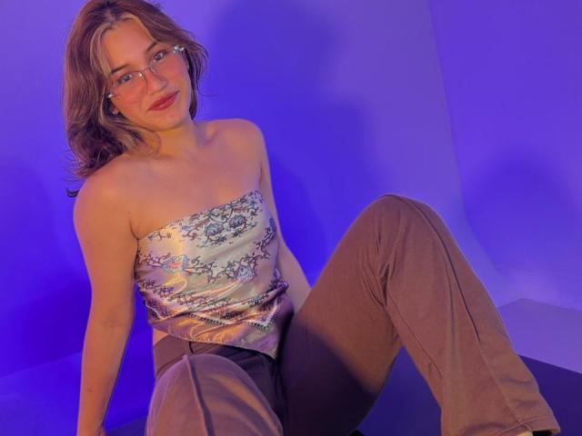 ElodieGardner with glasses, wavy brown hair, strapless silver top with floral pattern, brown pants, sitting, blue and purple lighting, smiling, medium tits, cute, relaxed, casual.