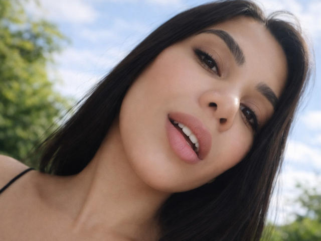 Anny, con el pelo largo y castaño oscuro, labios rosados ligeramente separados y ojos marrones, mirando hacia abajo con una sonrisa suave. Su rostro está centrado, con la cabeza ligeramente inclinada..