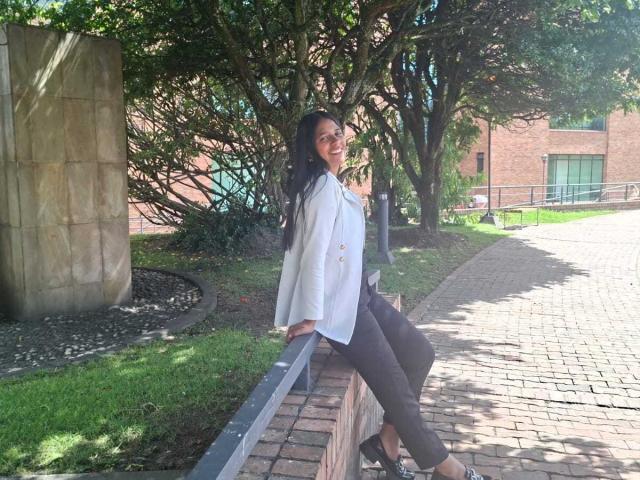 PauletteLoveX leans on a brick wall, wearing a white blouse, black pants, and black heels, smiling with dark curly hair, in a sunny outdoor setting.