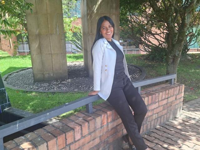 PauletteLoveX, staand, leunend tegen een bakstenen muur, gekleed in een witte blazer, een zwarte top en een broek, lachend, met lang zwart haar en zwarte hakken, in een zonnige buitenomgeving..