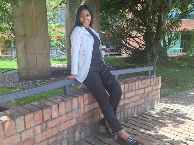 PauletteLoveX est appuyée contre un mur de briques, souriante, avec de longs cheveux noirs, une veste gris clair, un pantalon noir et des chaussures noires avec un motif. Ses fesses sont visibles..