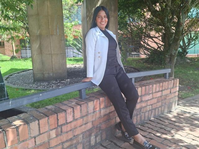 PauletteLoveX est une jeune femme latina adorable, aux cheveux foncés, assise sur un mur de briques, portant un blazer blanc, un haut noir et un pantalon, souriante, avec ses fesses appuyées contre le mur..