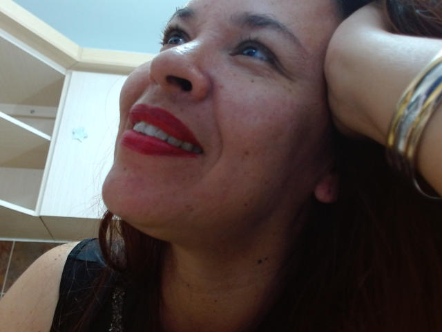 LizethBells has red lipstick, freckles, and is smiling with her head tilted up, showing her nose and chin. She's wearing a black top and gold bangles.
