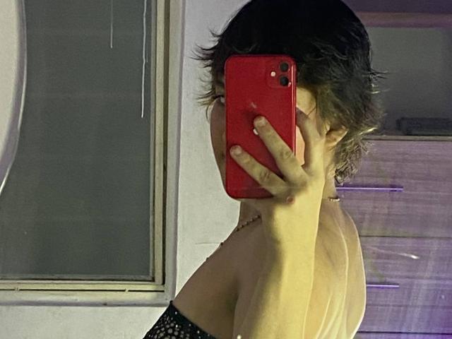 AdaraSweet with short, dark hair, holding a red phone, covering her face, showing her bare back and black strap, in a mirror selfie. Her hand's on her cheek.