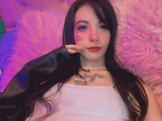 LaurenEvie69 con trucco rosa sul viso, giacca di pelle nera, canotta bianca, tatuaggi sul petto, capelli lunghi e scuri, piercing al naso, sdraiata su uno sfondo rosa e soffice, con una mano sul viso..
