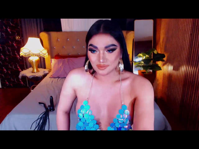 HottieBellias with blue sequin bra, showing off her big tits, in a bedroom with a lamp and bed in the background, looking directly at the camera with a pouty expression.