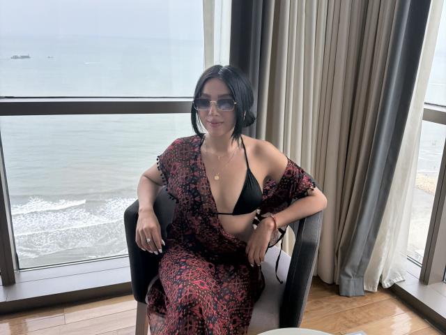 LillyTaylor with black bob, wearing a red and black patterned dress, black bikini top, glasses, sitting in a chair by a window, ocean view, wooden floor.
