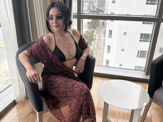 LillyTaylor, wearing a black bikini top and red patterned robe, sits in a chair with her tits showing, sunglasses on, smiling, next to a white table in a high-rise.
